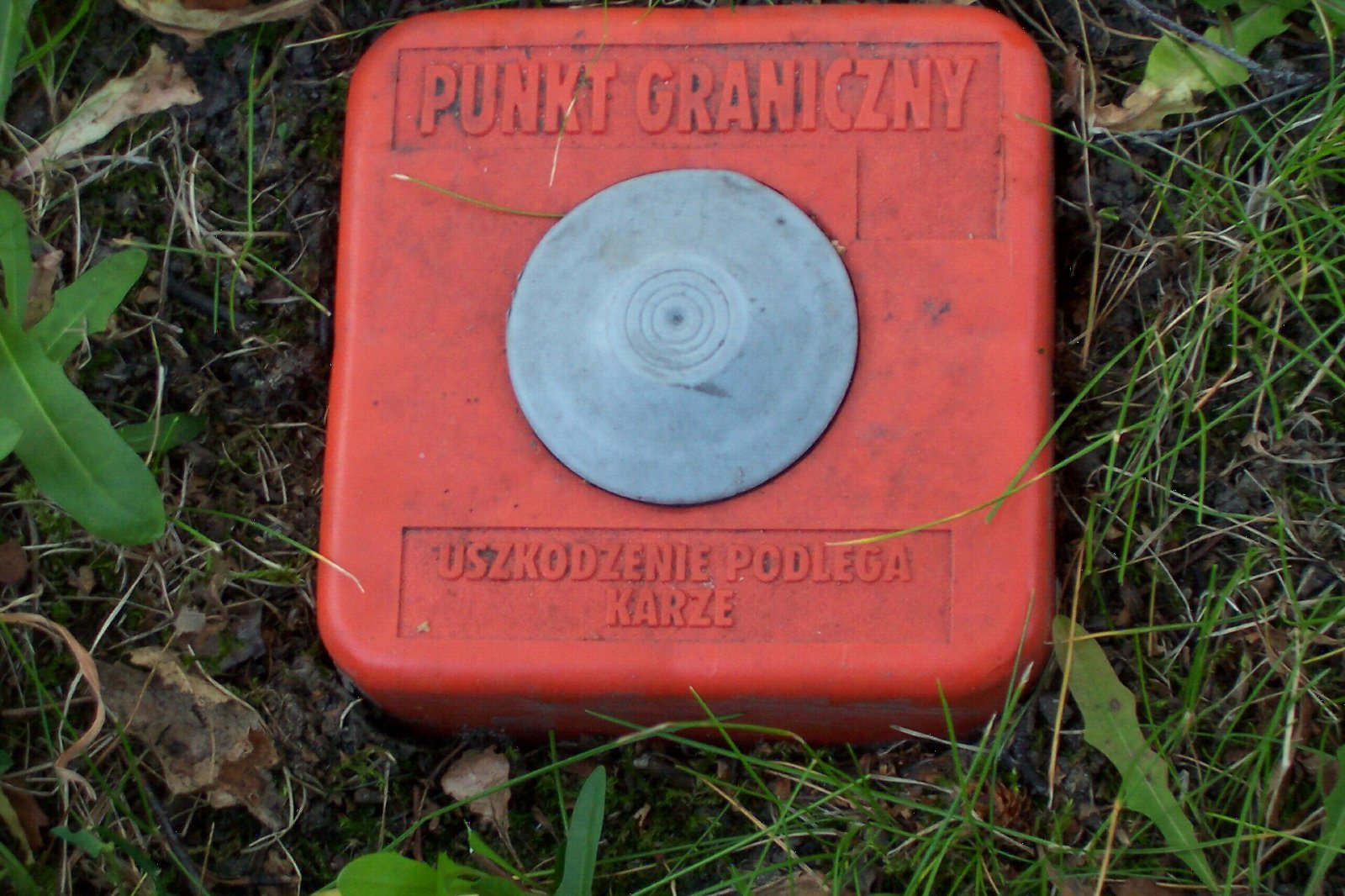 Pomarańczowy plastikowy znak z napisem Punkt graniczny i Uszkodzenie podlega karze, osadzony w trawie